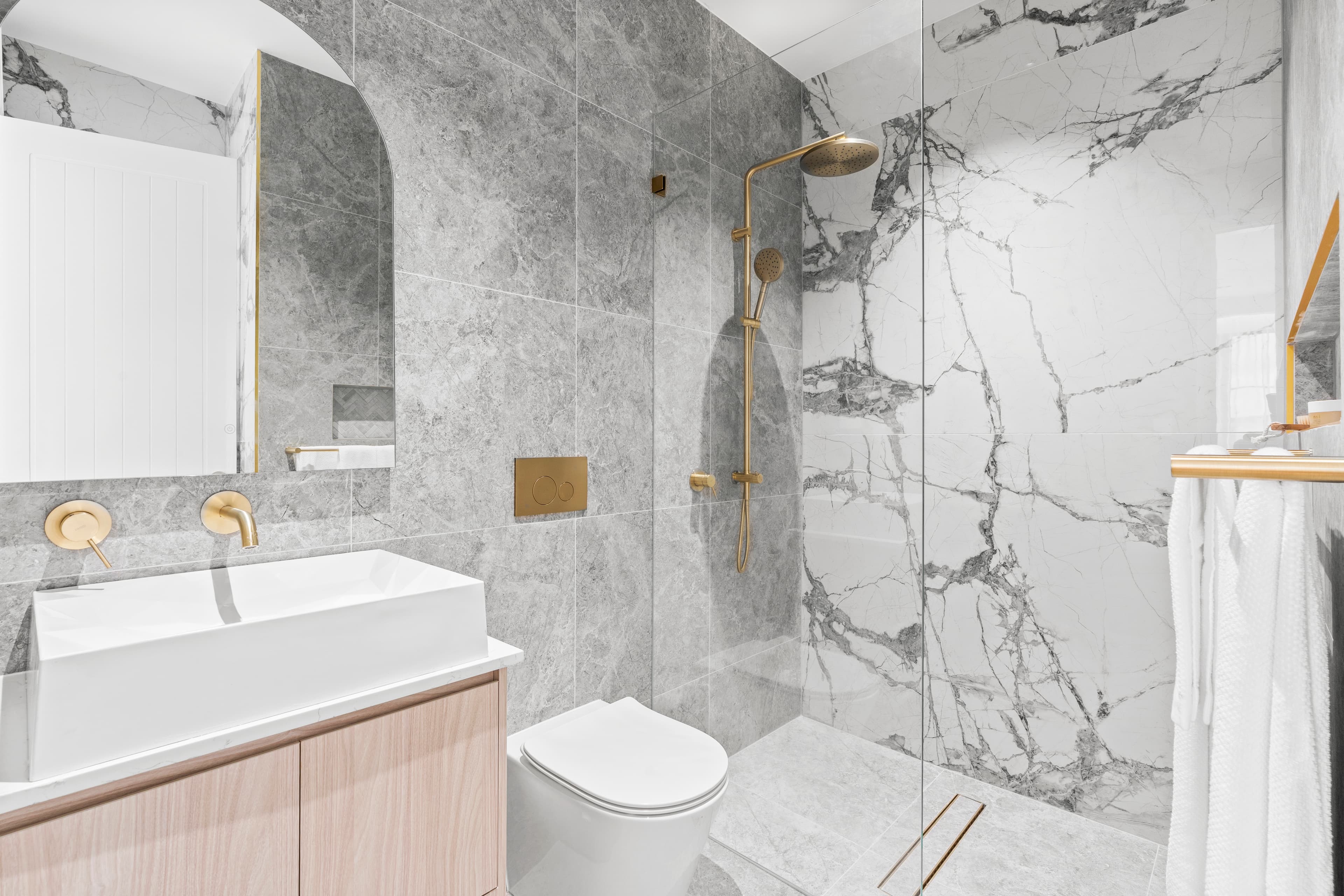 Compact ensuite with an arch mirror, grey marble feature wall and floating timber vanity by Reno Build on Sydney's Northern Beaches